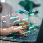Business Project Management System. Project manager working on laptop and updating tasks and milestones progress planning with Gantt chart scheduling interface for the company on virtual screen.